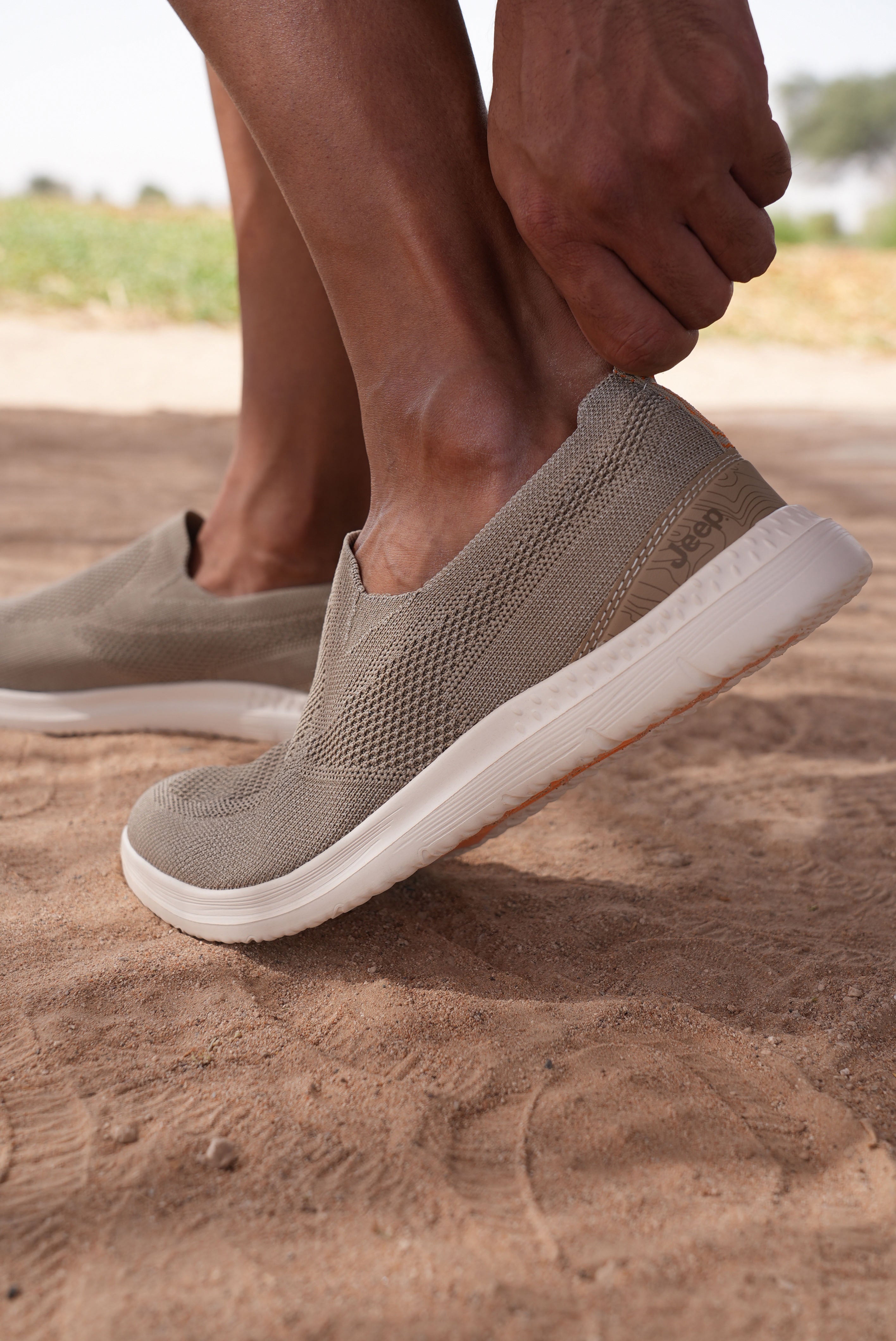 Man wearing beige slip-on Jeep shoes with white soles on a sandy surface.