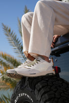 Man wearing off-white Jeep sneakers with green accents sitting on a Jeep tire, palm tree in the background