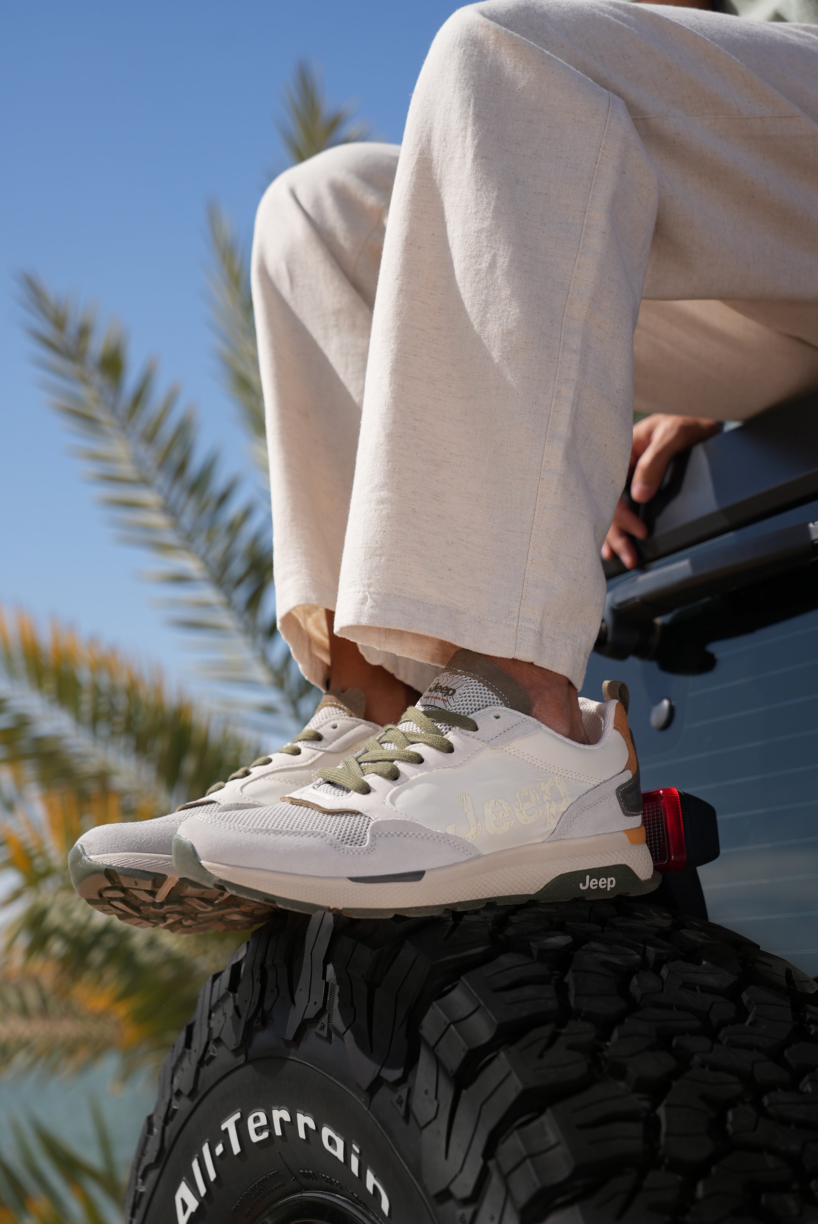 Man wearing off-white Jeep sneakers with green accents sitting on a Jeep tire, palm tree in the background