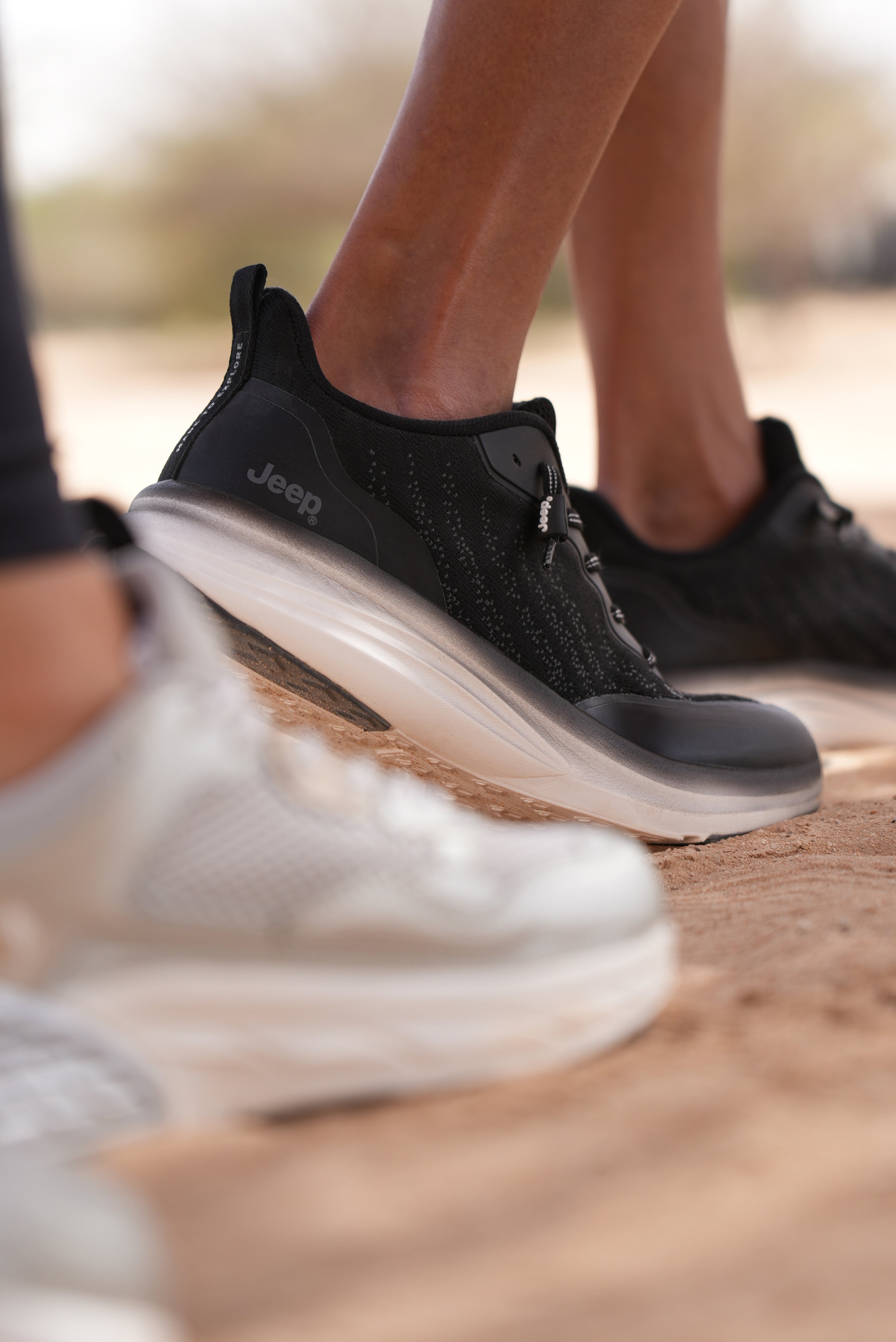 Black Jeep running shoes with white soles worn by a Man on a sandy background