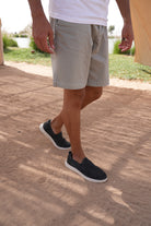 Man wearing beige shorts and black slip-on Jeep shoes on a sandy surface with grass and water in the background