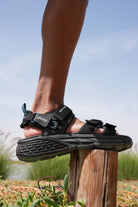 Man wearing black jeep hiking sandals with 'Jeep' logo on a wooden post against a natural background.